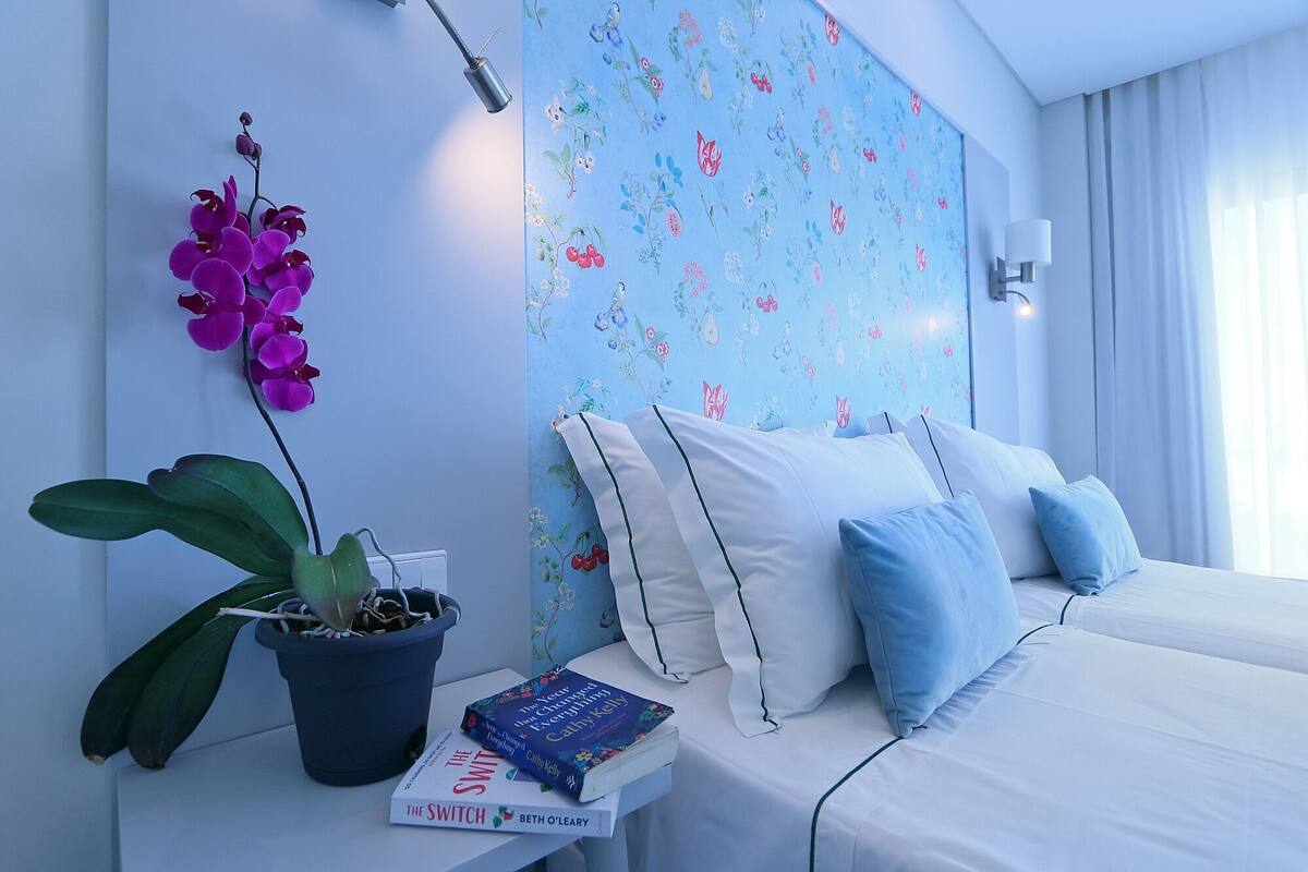 Une chambre d’hôtel avec un lit, une table de chevet, une plante en pot et un mur recouvert d’un papier peint à motifs floraux.