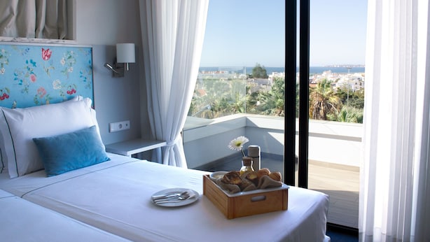 A hotel room with a bed, a tray with pastries, a plate with cutlery, and a view of the sea and buildings. - Agua Hotels Alvor Jardim (Portimão)
