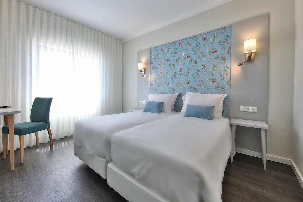 Een hotelkamer met twee bedden, een bureau, een stoel en een raam met gordijnen.