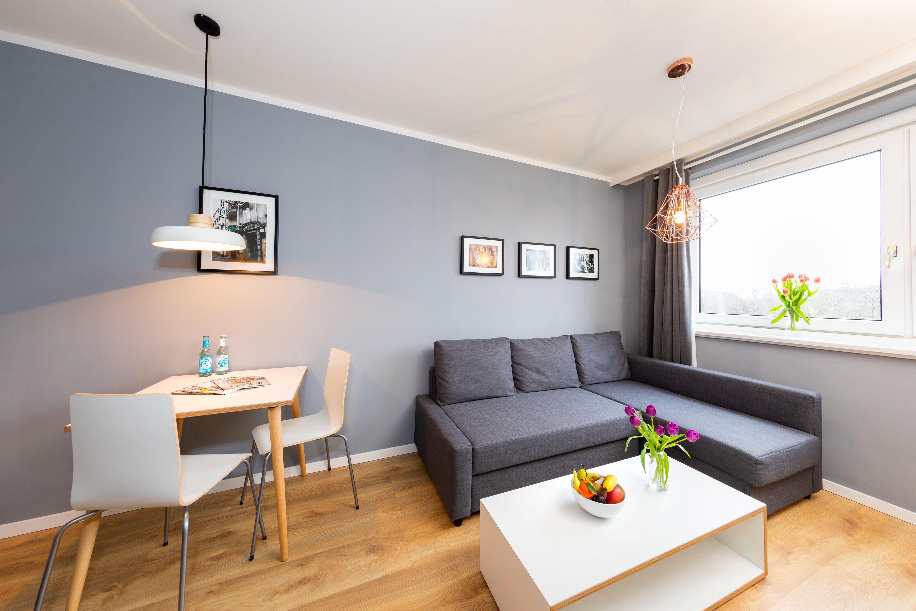 Un salon moderne avec un canapé gris, un fauteuil blanc, une table basse en bois et une table à manger avec deux chaises. On peut voir des tableaux encadrés au mur, une fenêtre avec vue et un luminaire suspendu.