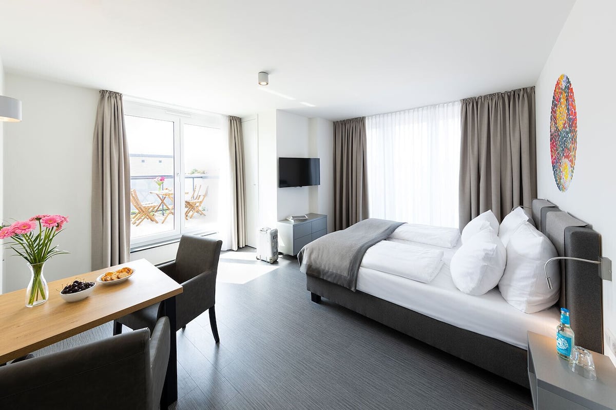 Una habitación de hotel moderna con cama, escritorio, silla, televisión y balcón con mobiliario exterior.