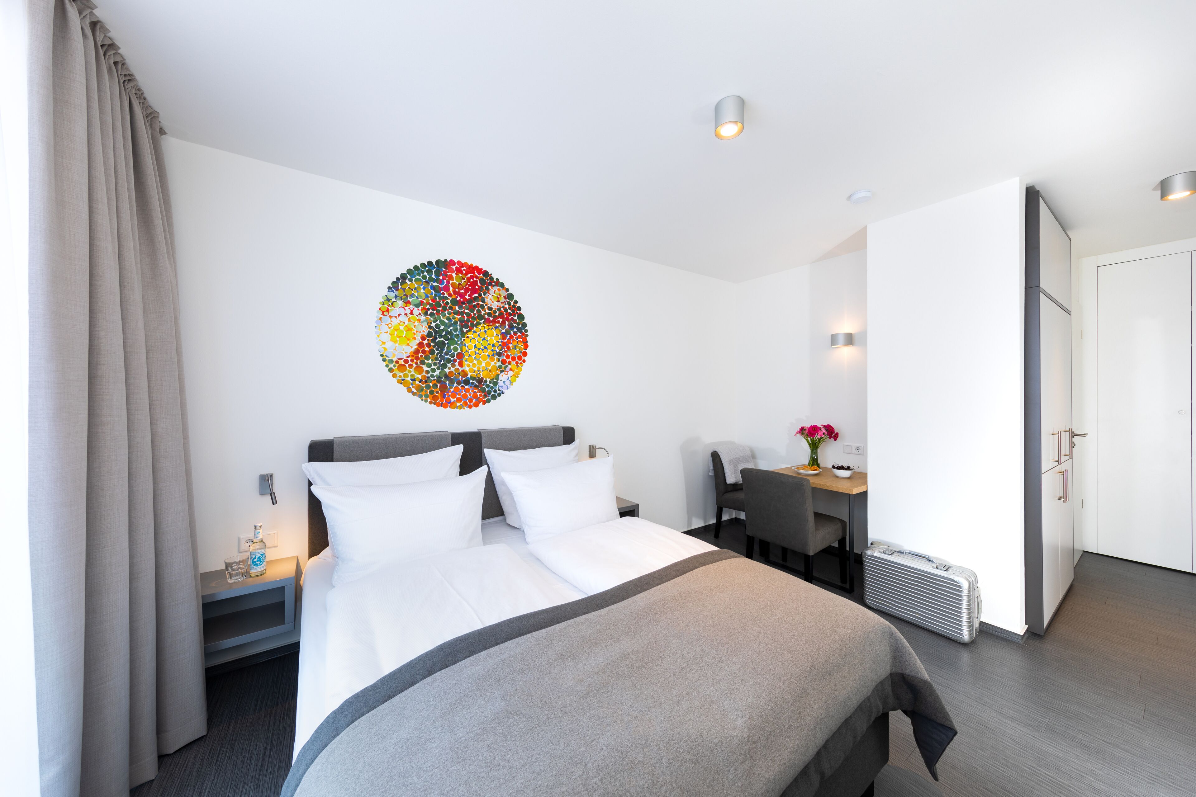 A modern hotel room with a neatly made bed, a desk with a chair, a small table with a vase of flowers, and a radiator.
