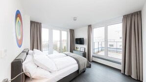 Habitación de hotel moderna con una cama grande, un televisor de pantalla plana, una mesita de noche y vistas de la ciudad a través de amplios ventanales.