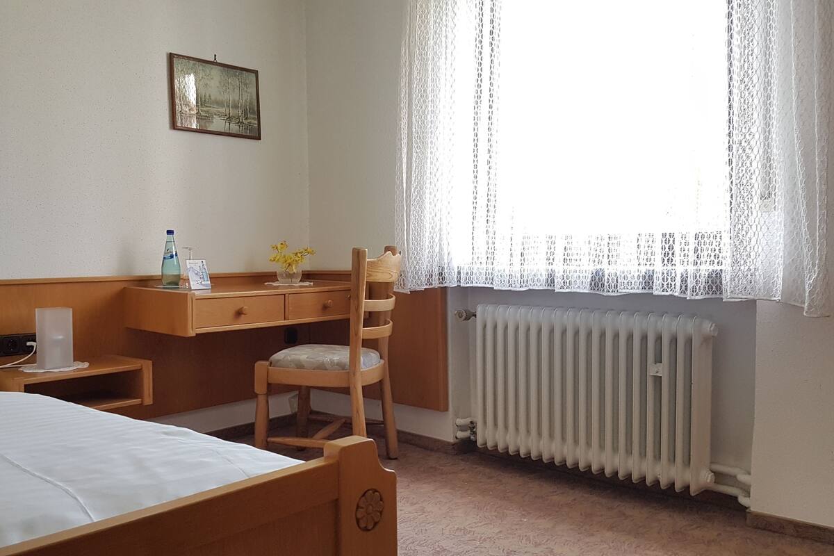 Een hotelkamer met een bed, een houten bureau, een stoel, een radiator en een raam met kantgordijnen.