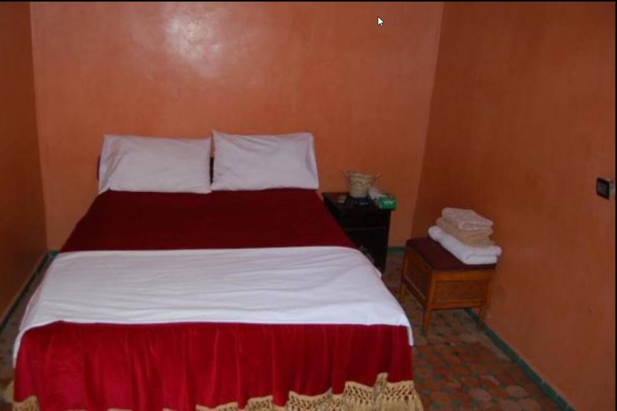 A bed with a red and white bedspread, a nightstand with a lamp, and a small table with towels.