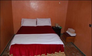 A bed with a red and white bedspread, a nightstand with a lamp, and a small table with towels. - Hotel Jardin Public (Fes)