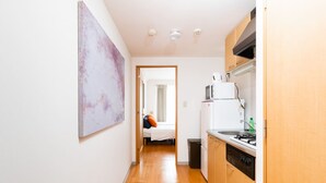 A narrow hallway with a wooden floor leading to a bedroom with a bed and a window. A kitchen area is visible on the right with a stove, microwave, and sink. - Guest House MIYABI (Kyoto)