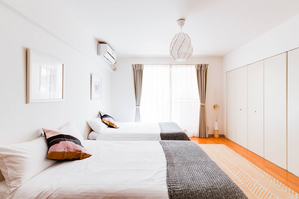 Quarto de hotel com duas camas, uma janela grande com cortinas, um guarda-roupa e um candeeiro pendente.