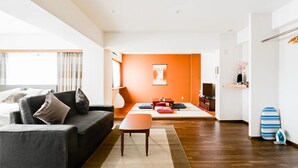 A modern living room with a sofa, a coffee table, and a TV. There is a wooden floor and a bright orange accent wall. - Guest House MIYABI (Kyoto)