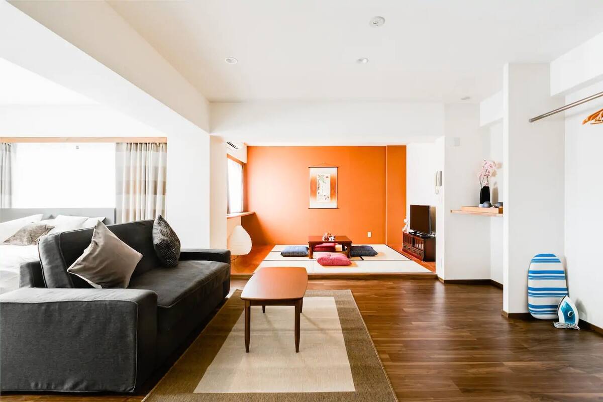 Uma sala de estar moderna com um sofá, uma mesa de centro e uma televisão. O chão é de madeira e há uma parede de destaque laranja vibrante.