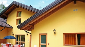 A yellow building with a wooden balcony, a door with a window, and a table with chairs and an umbrella. - Garnì Sella al Cipriani (Borgo Valsugana)