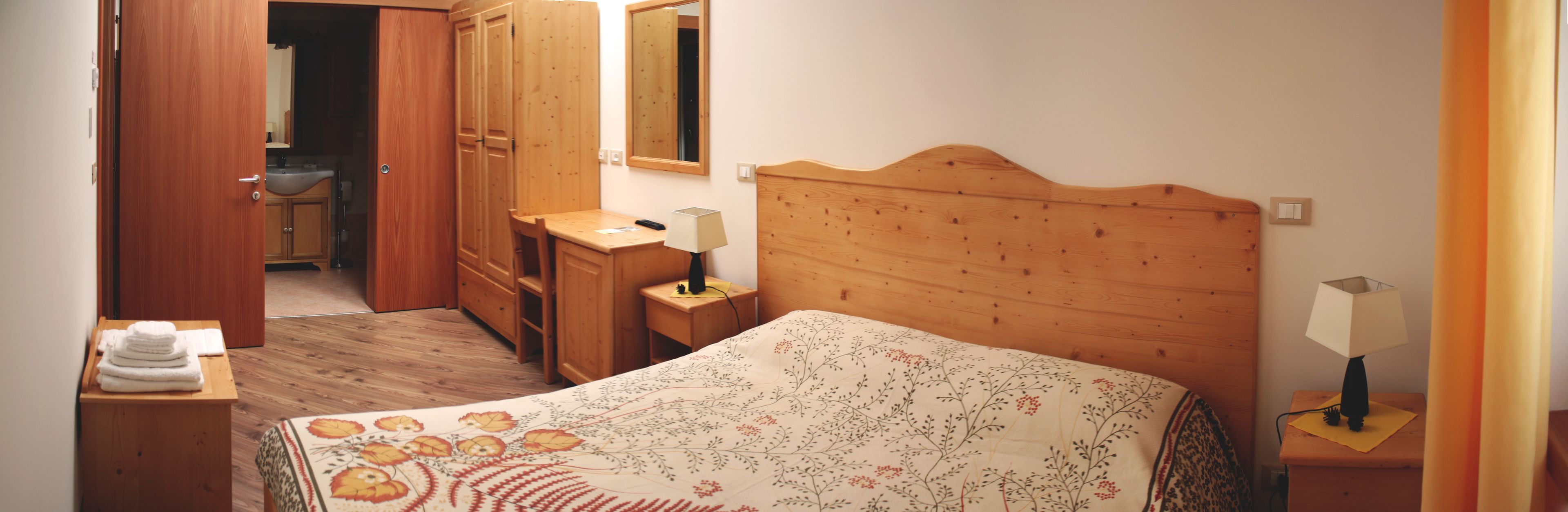 A bedroom with a wooden headboard, a bed with a floral bedspread, a bedside table with a lamp, a wooden dresser, and a mirror.