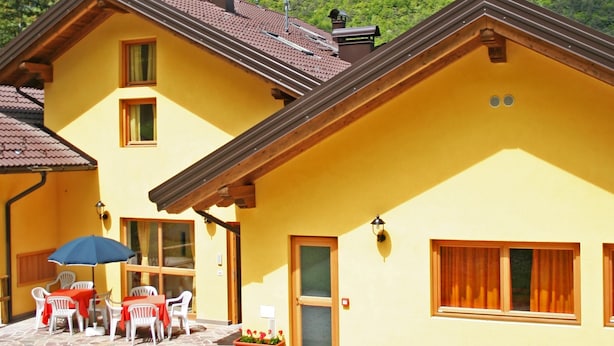 A yellow building with a tiled roof, a patio with outdoor furniture, and a mountainous backdrop. - Garnì Sella al Cipriani (Borgo Valsugana)