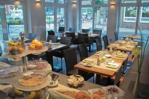 A dining area with multiple tables set for meals, a buffet with various food items, and large windows providing a view of the outside. - HIB Hotel in Baesweiler (Baesweiler)
