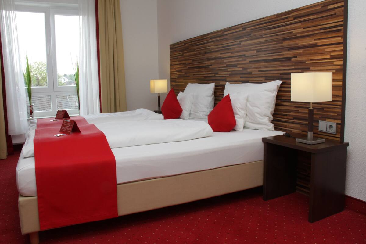 Un lit double avec du linge de lit blanc et des touches de rouge, une tête de lit en bois, deux tables de chevet avec des lampes et un sol recouvert d’une moquette rouge.