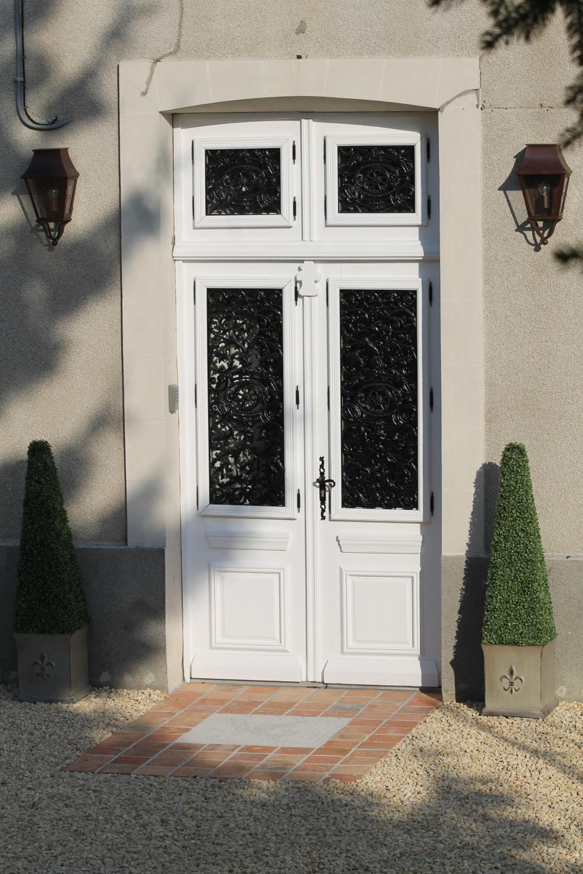 A double door with decorative glass panels and a white frame, flanked by two wall-mounted lanterns and two topiary bushes.