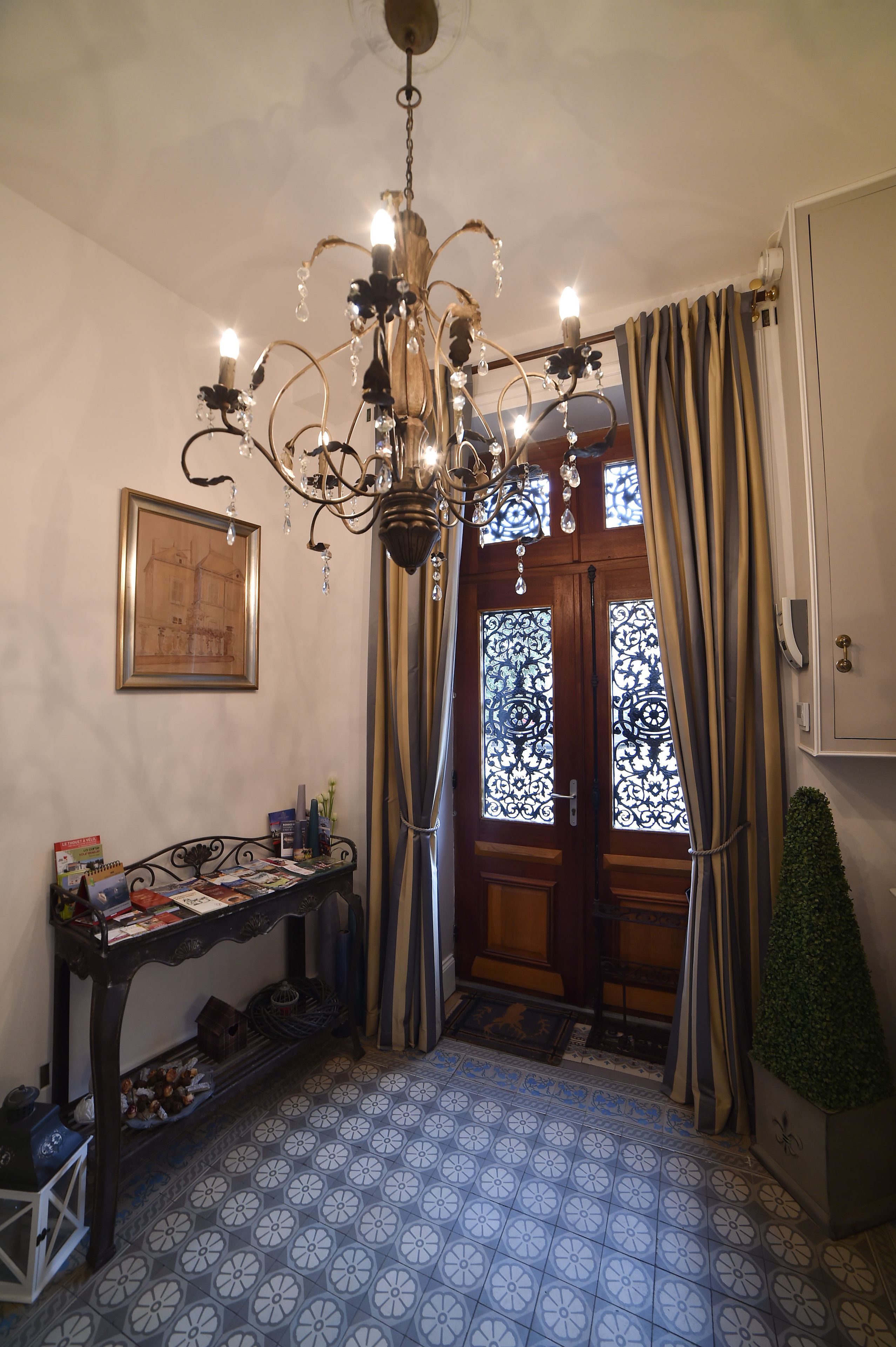 A room with a chandelier, a wooden door with decorative glass panels, a table with books and a small plant, and patterned floor tiles.