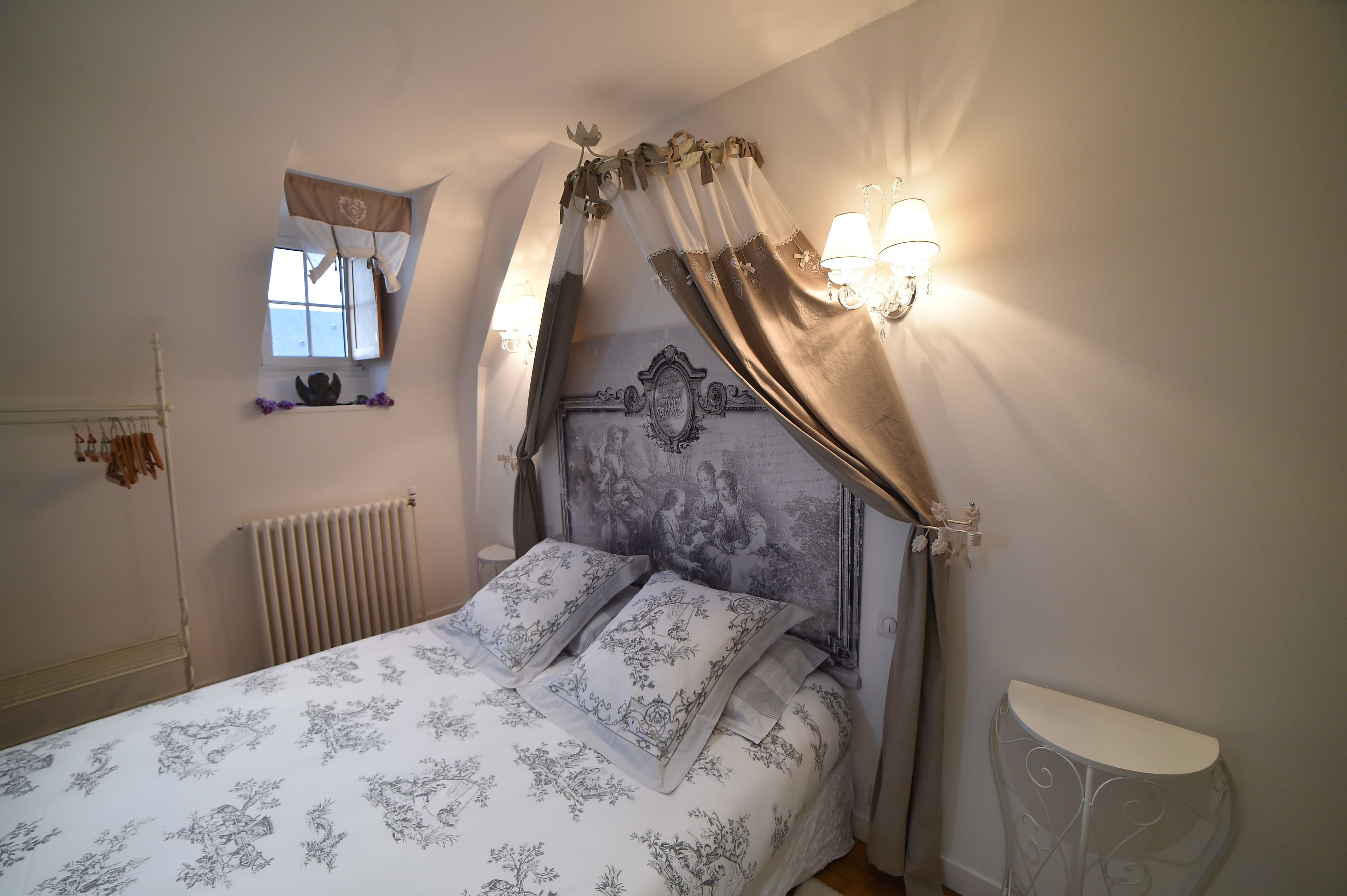 A bedroom with a bed, a nightstand, a wall-mounted lamp, a radiator, and a window with curtains.