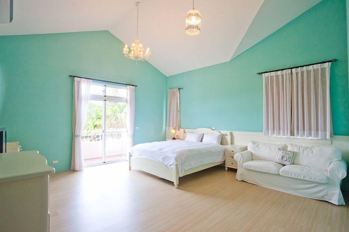 A bedroom with a bed, a white sofa, a chandelier, and a window with curtains.