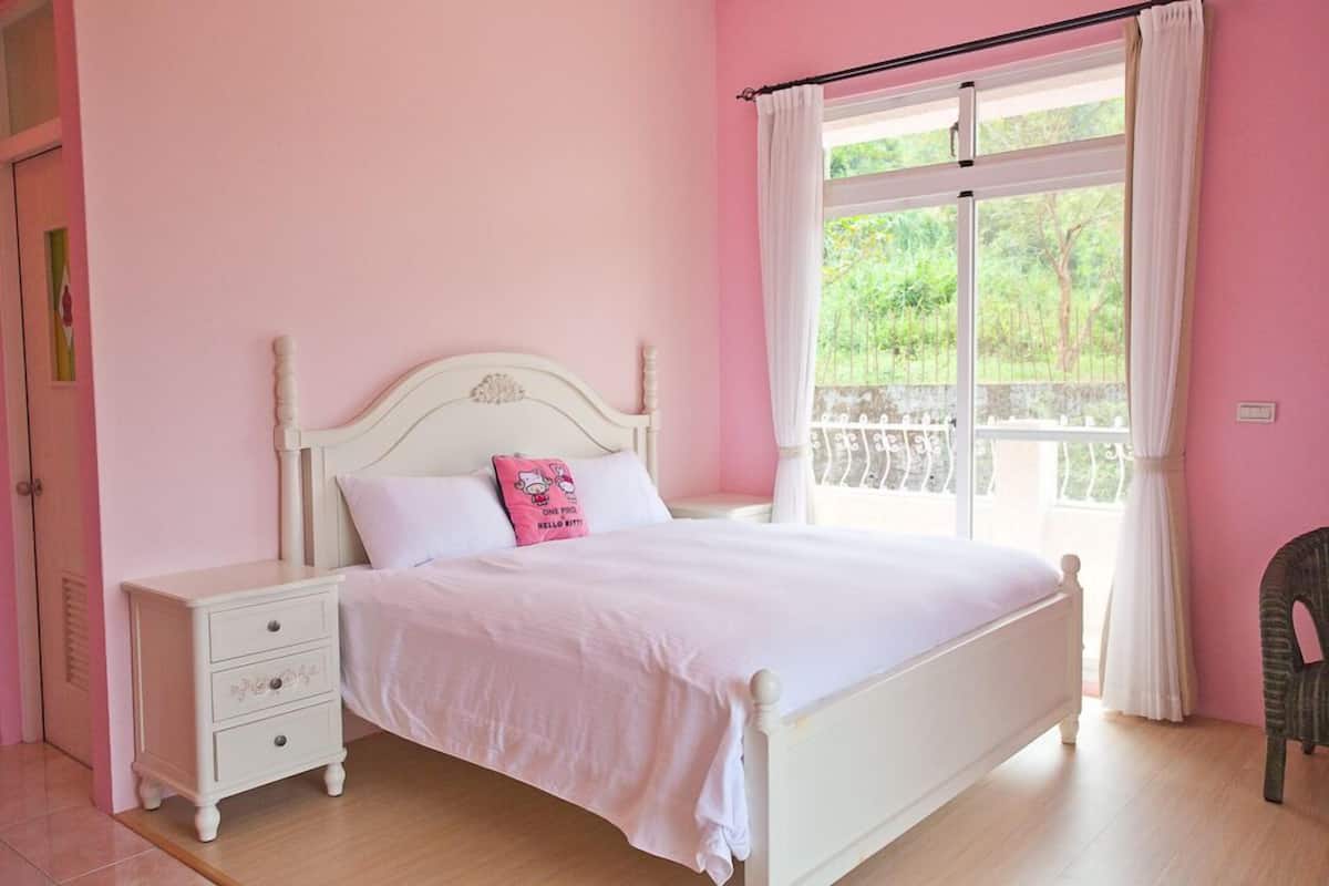 A bedroom with a white bed, a nightstand, a window with curtains, and a view of greenery outside.