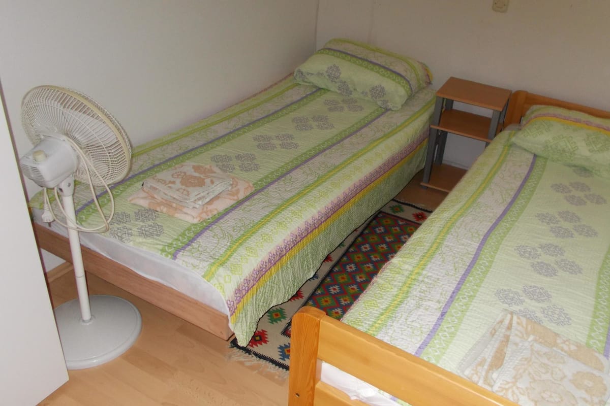 Eine kleine, einfache Unterkunft mit zwei Einzelbetten, einem Standventilator und Holzboden.
