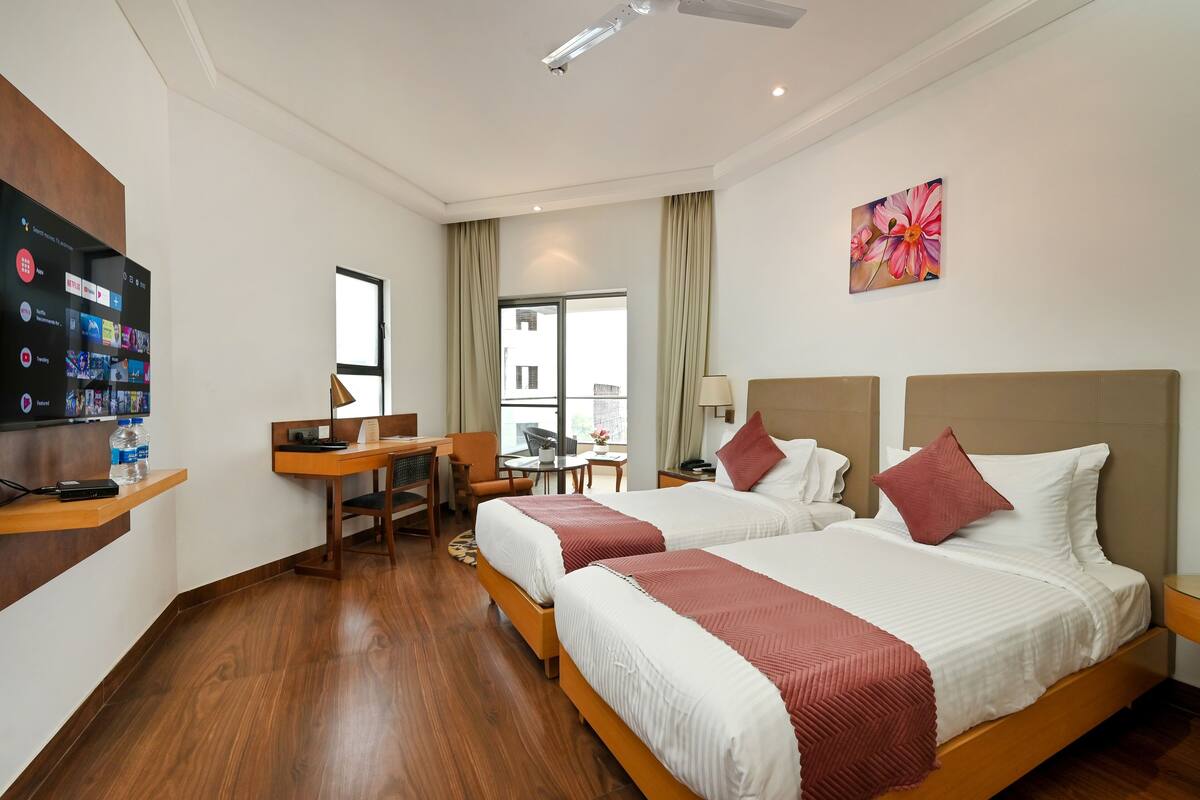 A hotel room with two beds, a desk, a chair, a TV, and a window.