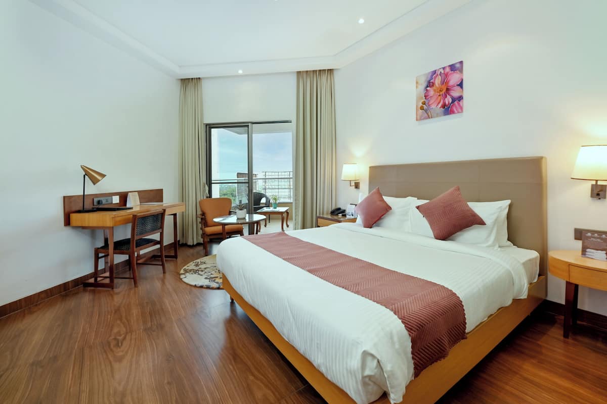 A hotel room with a large bed, a desk, a chair, a small table, and a balcony with a view.