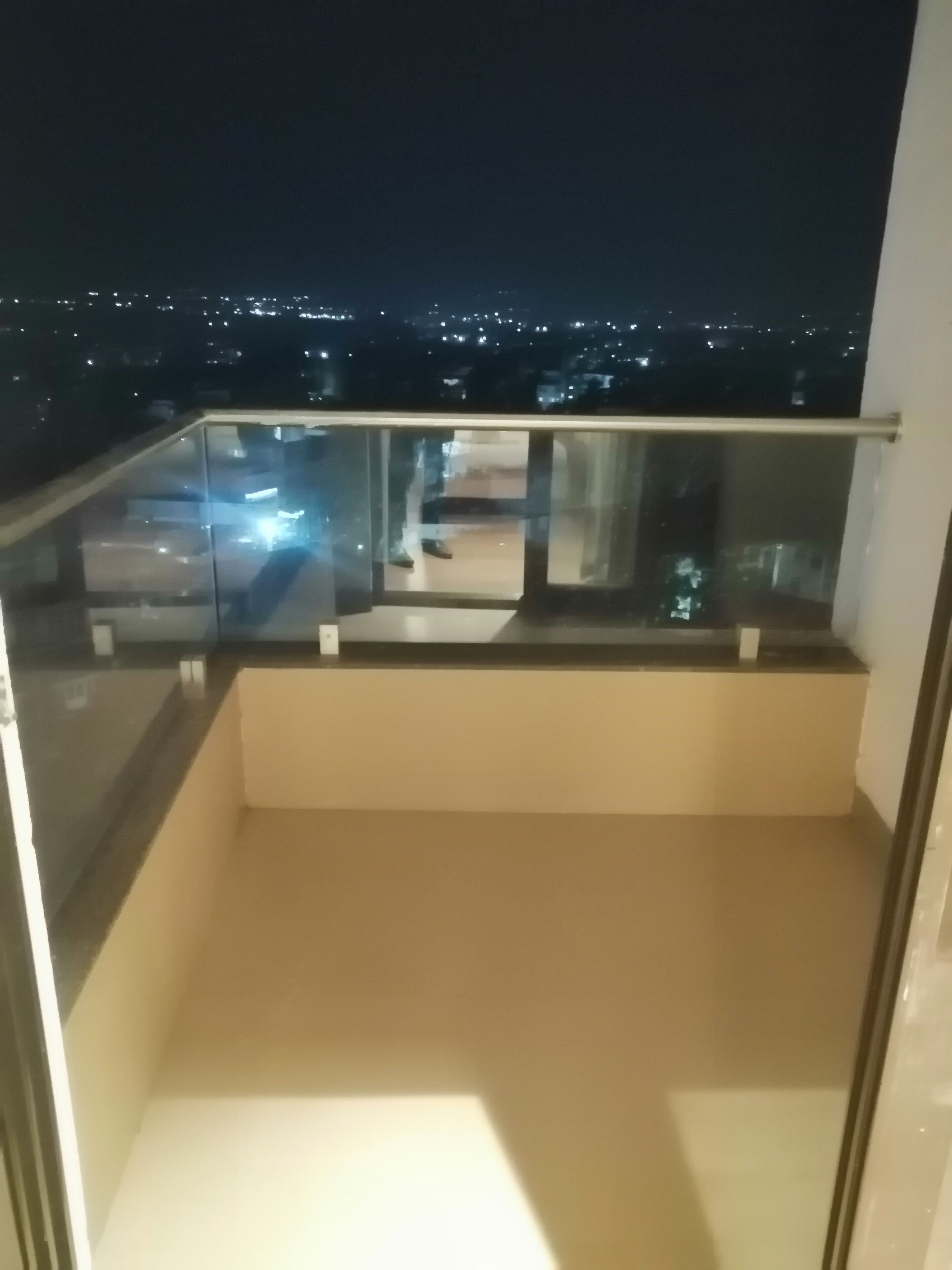 A balcony with a glass railing, a view of a cityscape at night, and a flat roof.