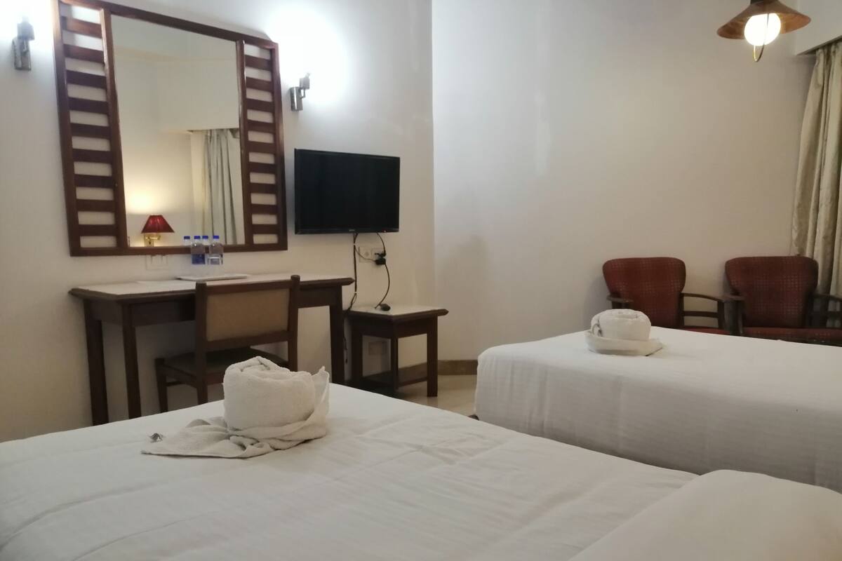 A hotel room with two beds, a desk, a chair, a TV, and a ceiling fan.