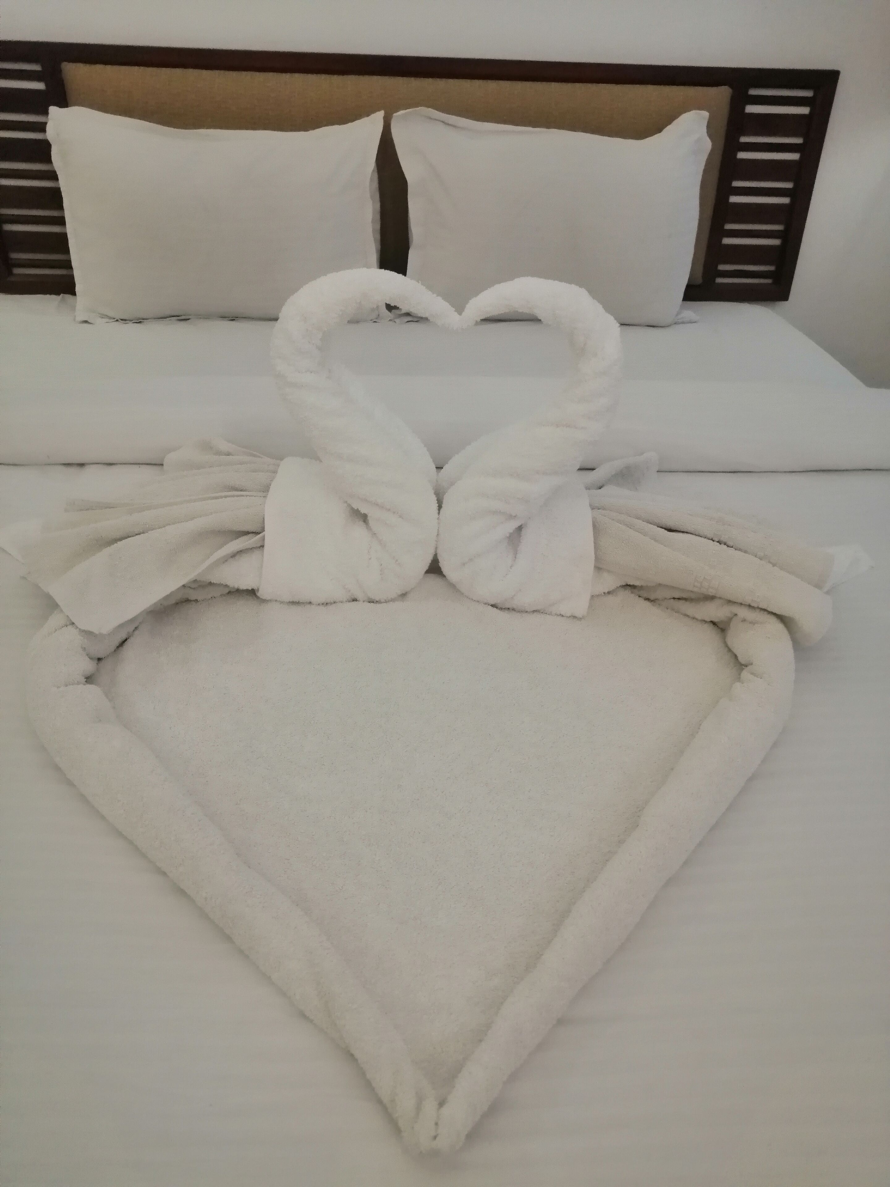 A neatly made bed with white linens, a heart-shaped towel arrangement, and a headboard with a wooden frame.
