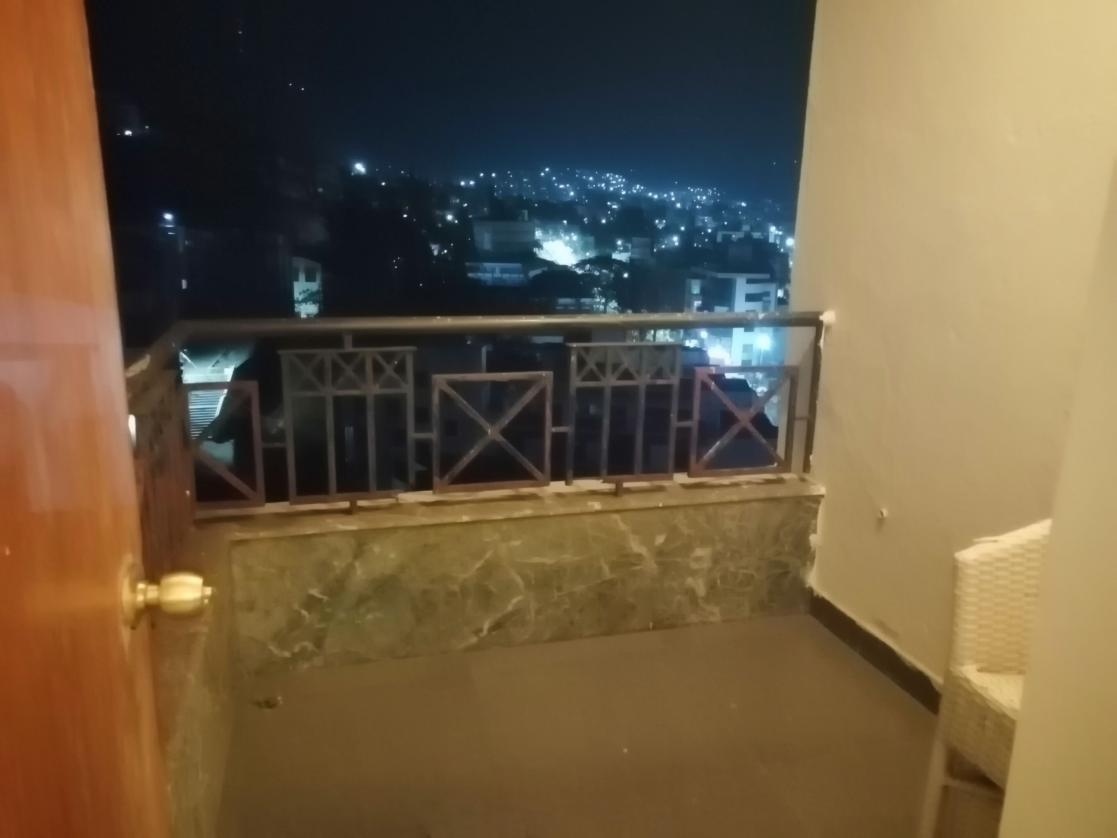 A balcony with a metal railing, a stone floor, and a view of a cityscape at night.