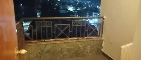 A balcony with a metal railing, a stone floor, and a view of a cityscape at night.