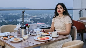 A woman sitting at a table with a city view, a coffee pot, and various food items. - Maharaja Regency (Satara)