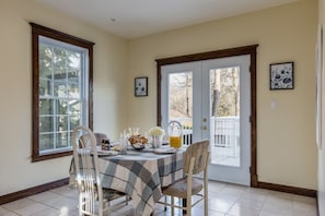 A dining area with a table set for breakfast, including bread, fruit, and a pitcher of orange juice. There are two chairs, a window, and a door leading outside.