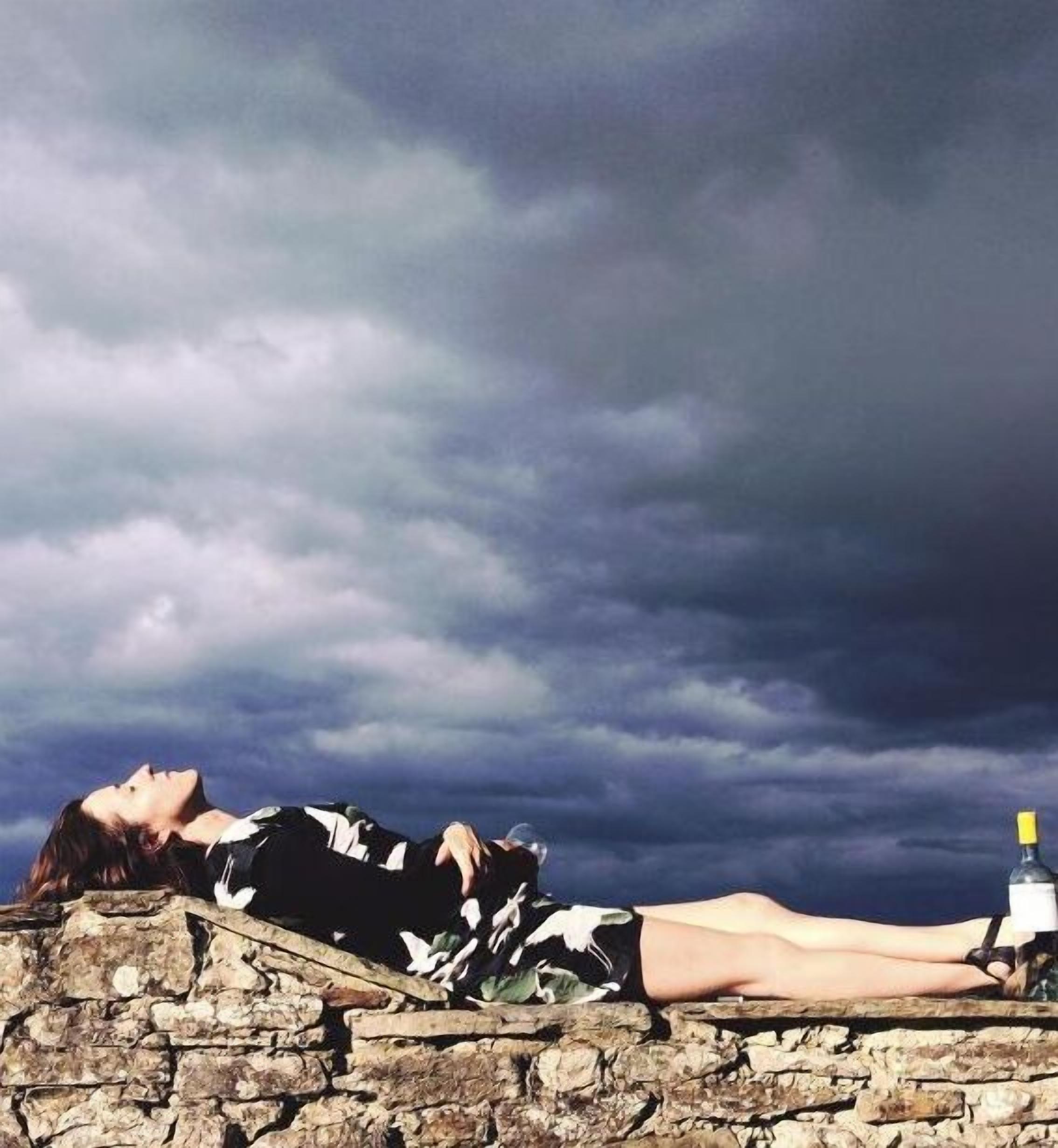 A person lying on a stone wall with a bottle and a bird figurine beside them, under a cloudy sky.