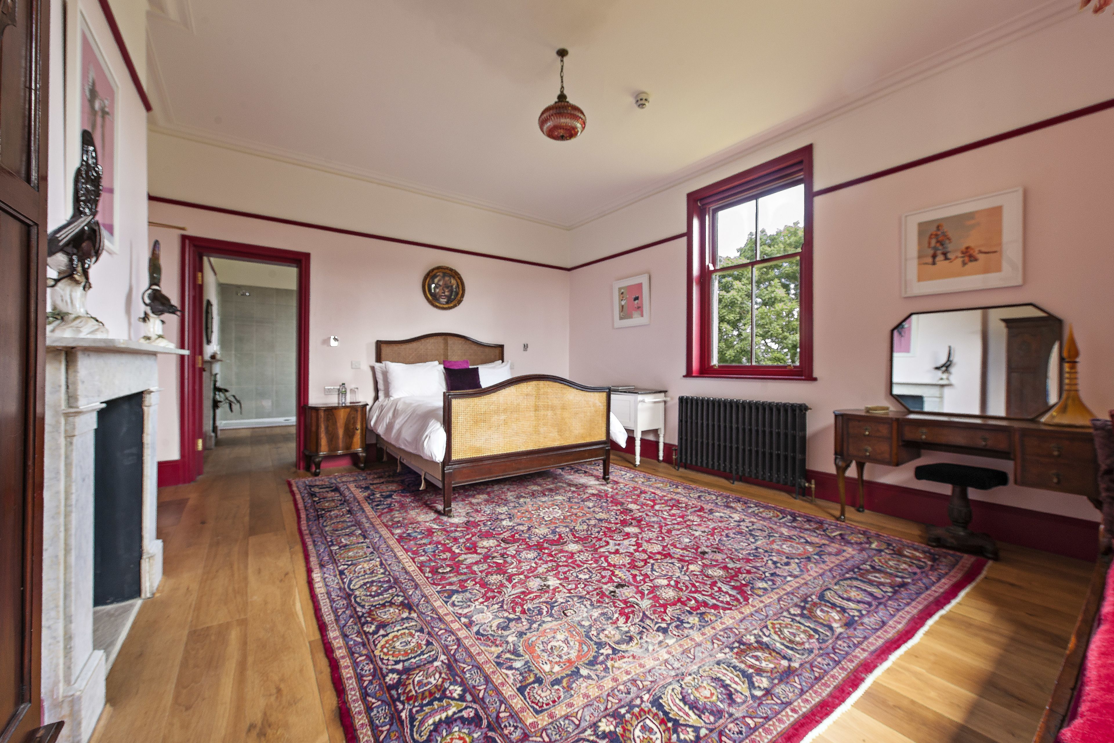 Un dormitorio con un tapete grande y ornamentado, una cama de madera, un aparador, un espejo y una ventana con vistas a árboles.