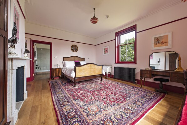 A bedroom with a large ornate rug, a wooden bed, a dresser, a mirror, and a window with a view of trees.