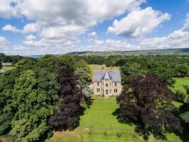 Um edifício grande e histórico com fachada simétrica, rodeado por vegetação exuberante e um gramado bem cuidado.