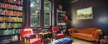 Quarto com uma grande estante de livros, uma poltrona vermelha, um banquinho azul e um candeeiro vintage.