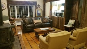 A cozy living room with a leather sofa, wooden coffee table, and two armchairs. A vintage stove is on the left, and a window with curtains is visible. - 1860 House Inn and Vacation Rental Home (Stowe)