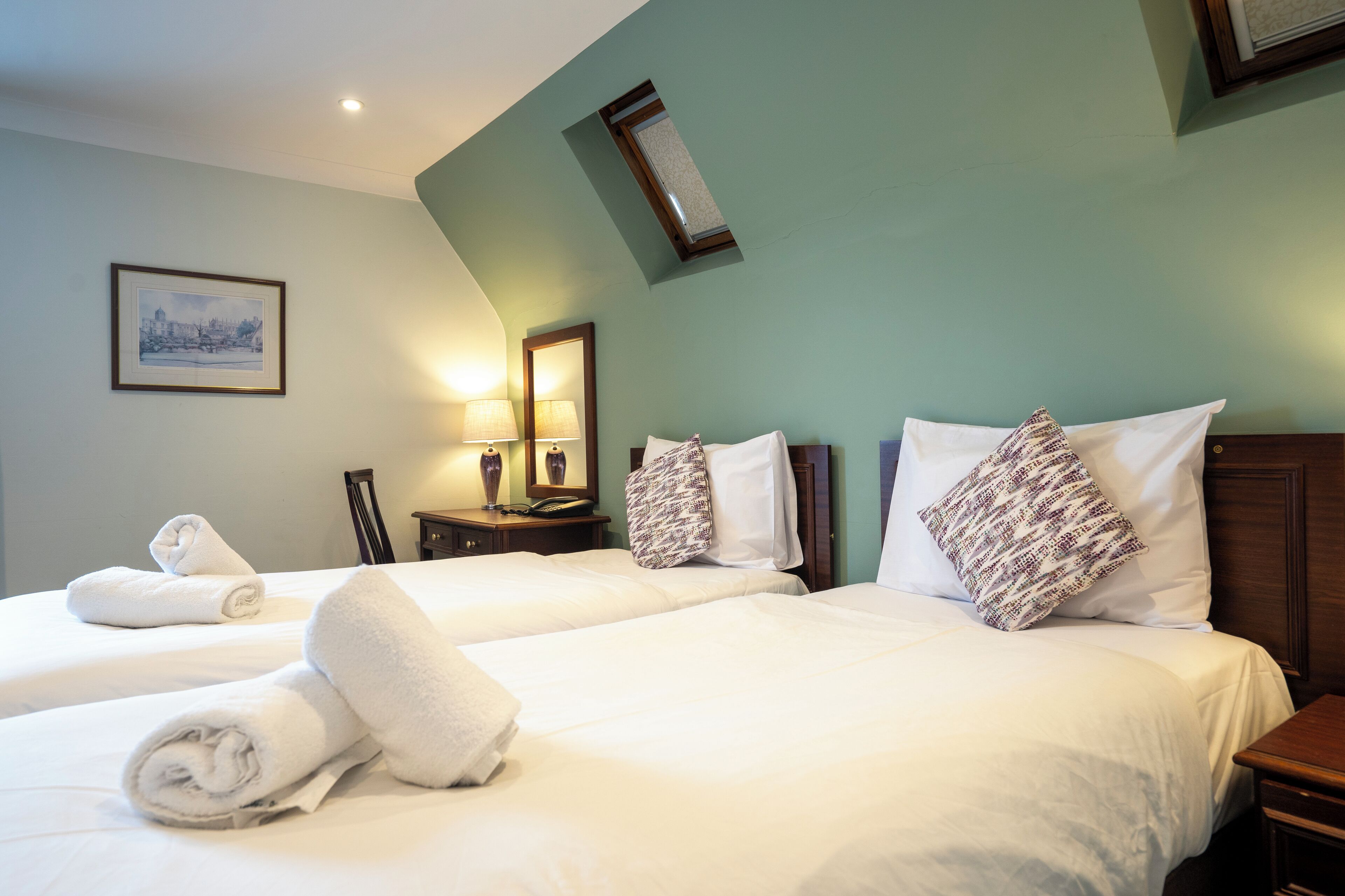 A hotel room with two beds, white linens, and towels. There is a framed picture on the wall and a small table with a lamp.