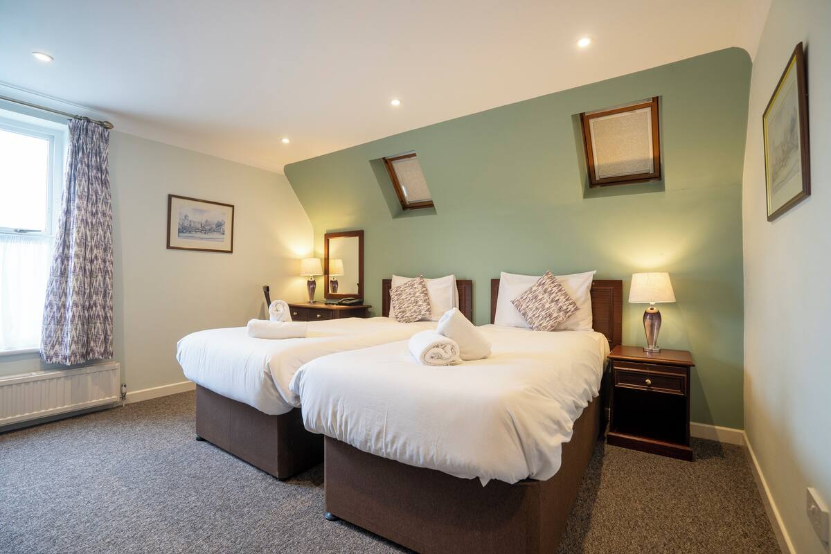 A hotel room with two beds, a window with curtains, a radiator, and a nightstand with a lamp.