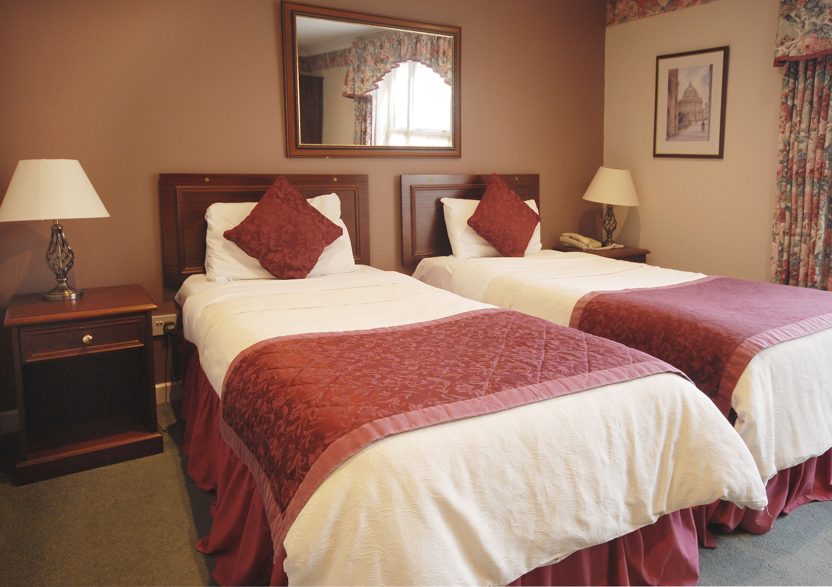 Habitación de hotel con dos camas, cada una con colcha roja y ropa de cama blanca. Hay dos mesitas de noche con lámparas, un espejo sobre las camas y un cuadro enmarcado en la pared.