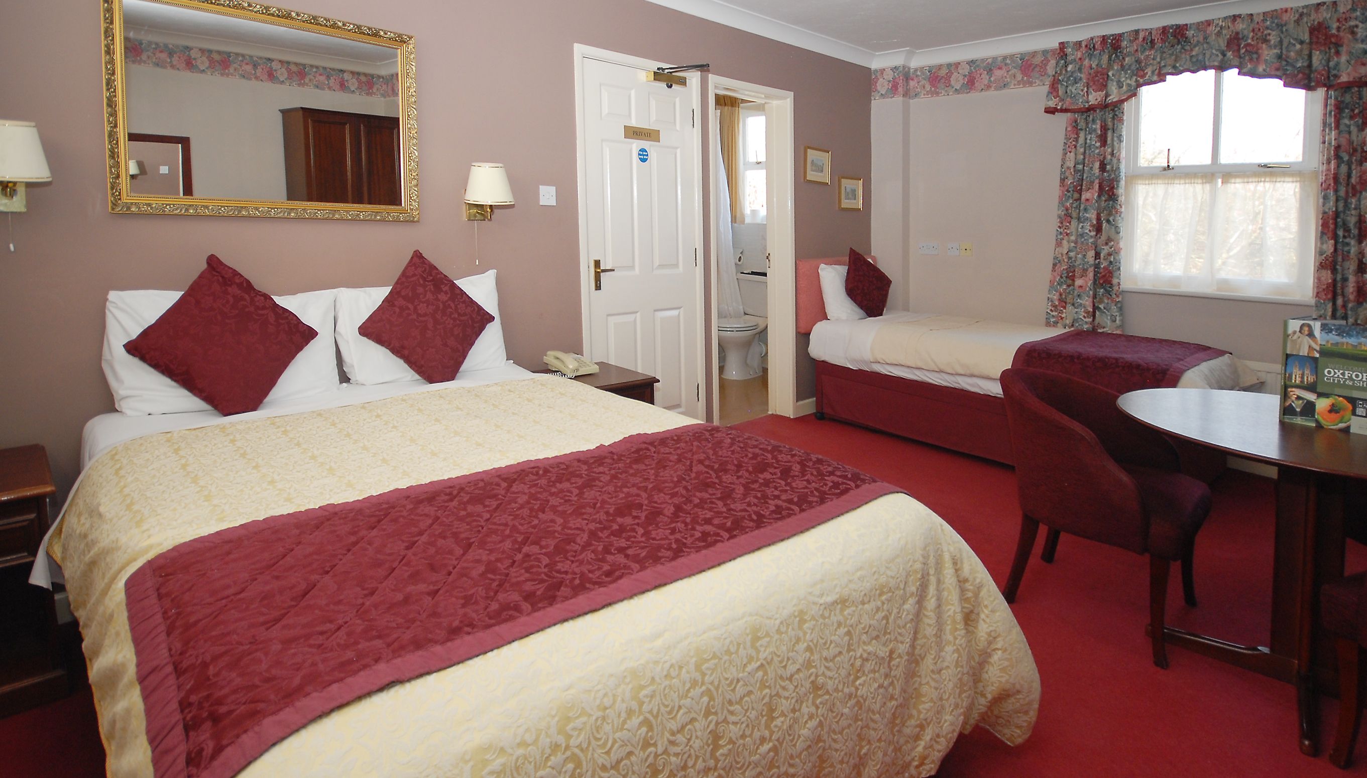A hotel room with two beds, a desk, a chair, a mirror, and a window with floral curtains.