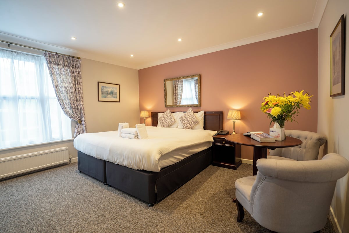 A hotel room with a large bed, bedside lamps, a chair, a small table with a vase of flowers, and a window with curtains.