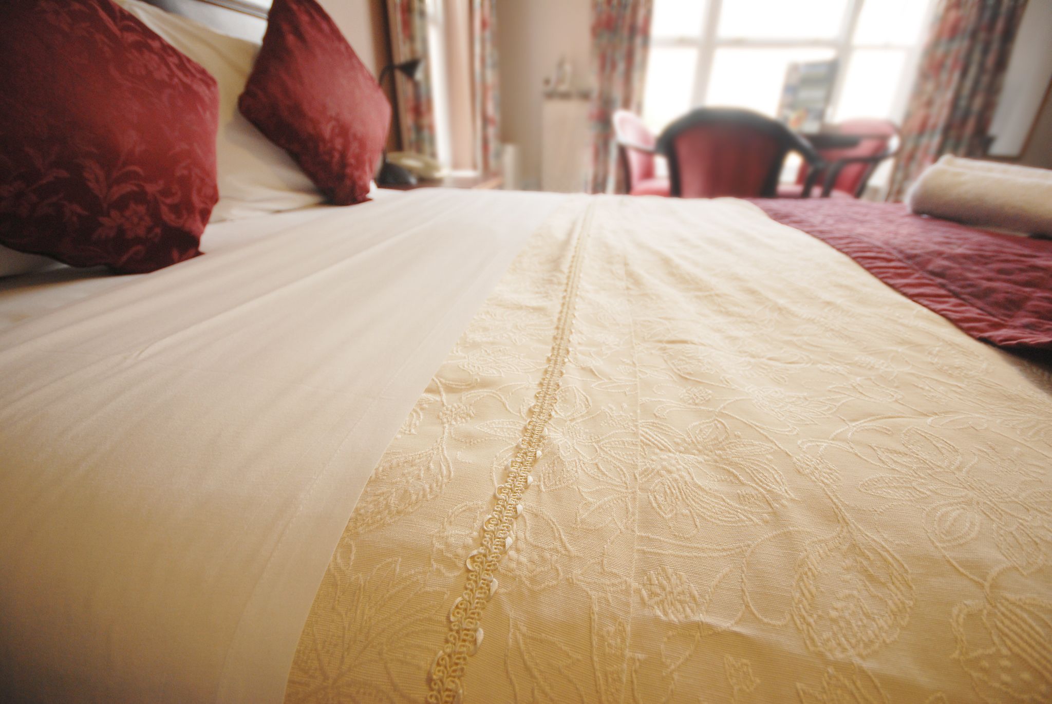 A bed with a white and beige bedspread, red pillows, and a chair in the background.
