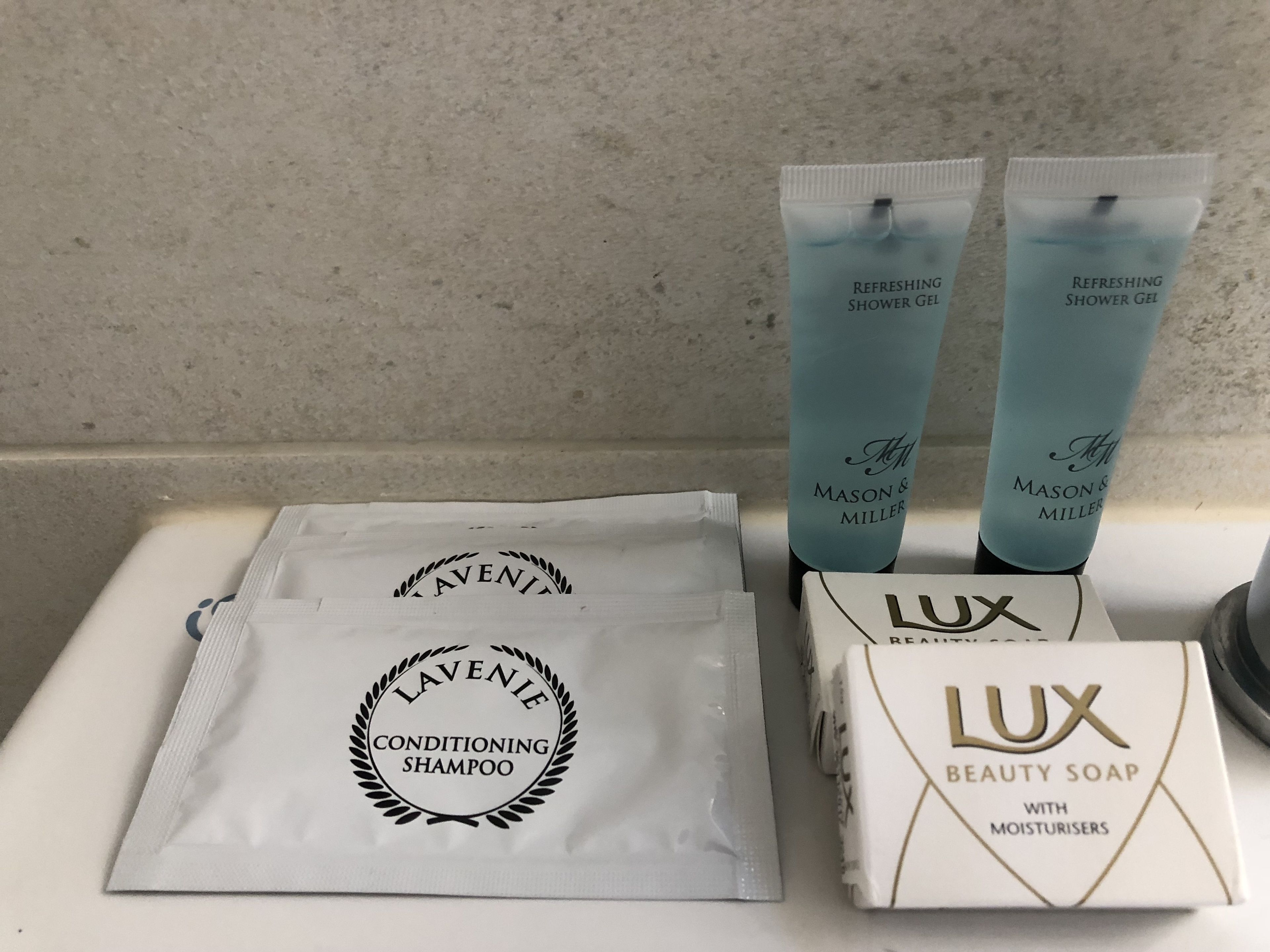 Una encimera de baño con artículos de aseo, incluyendo champú acondicionador LAVENIE, dos tubos de gel de ducha refrescante MASON & MILLER y una pastilla de jabón LUX con agentes hidratantes.