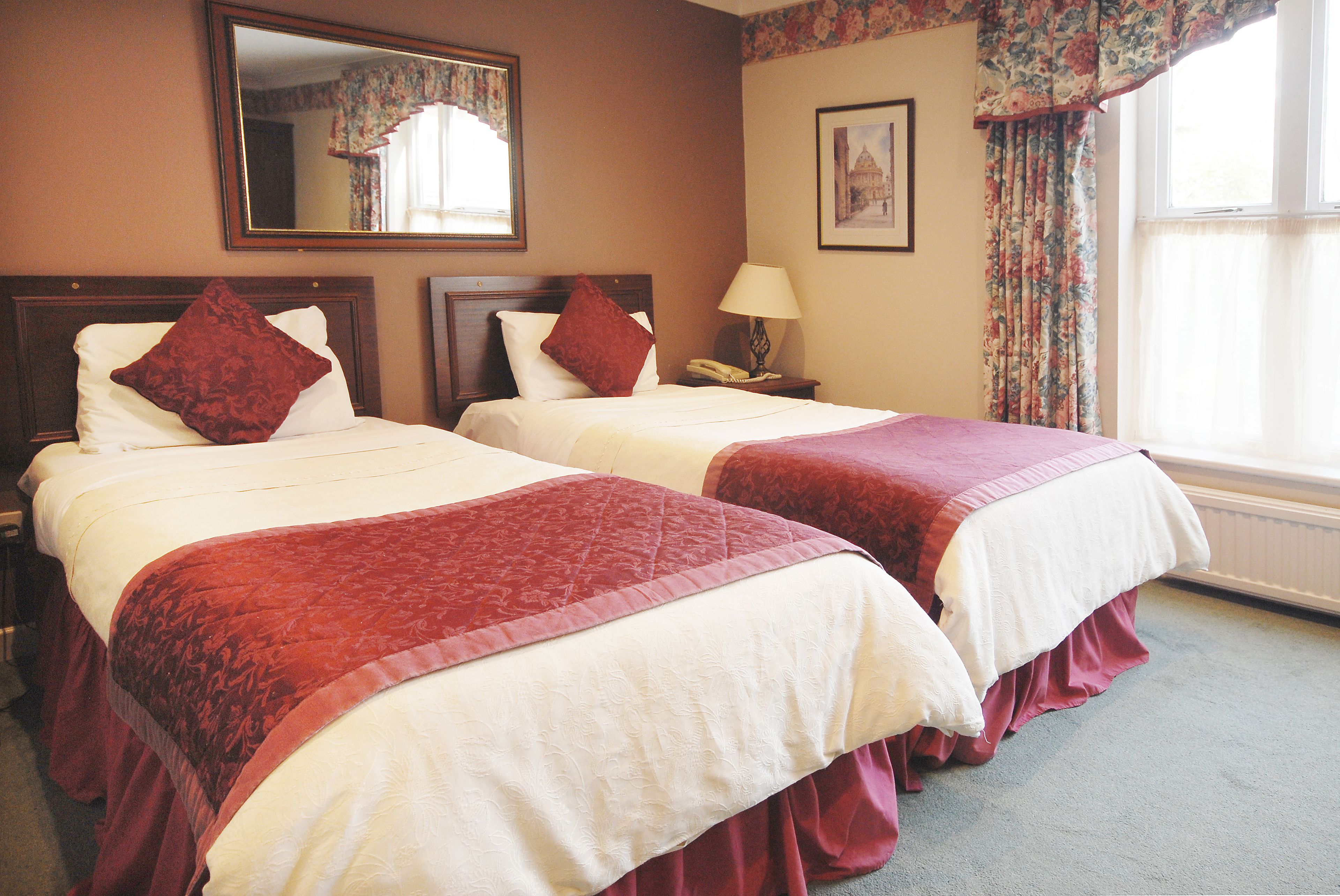 A hotel room with two beds, each with a red bedspread and white pillows. There is a nightstand with a lamp and a telephone, a mirror on the wall, and a framed picture above the bed.