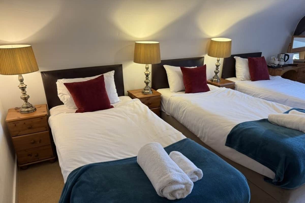 A hotel room with two beds, each with a white comforter and red pillows. There are two bedside tables with lamps and a small wooden dresser.