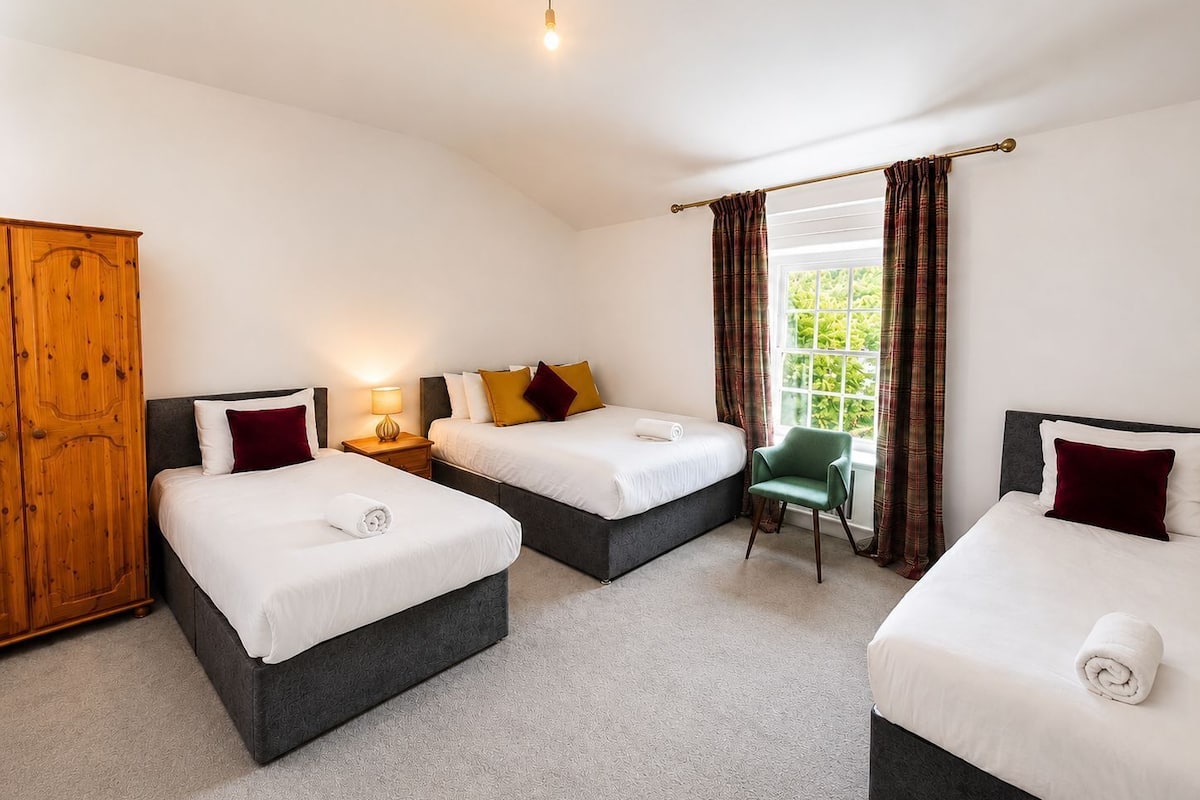 A hotel room with three beds, a wooden wardrobe, a green chair, and a window with curtains.